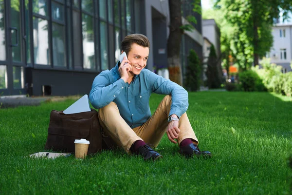 Handsome smiling young male freelancer sitting on grass and talking by smartphone — Stock Photo