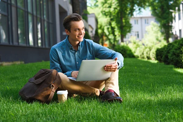 Schöner lächelnder junger Freiberufler, der auf Gras sitzt und Laptop benutzt — Stockfoto