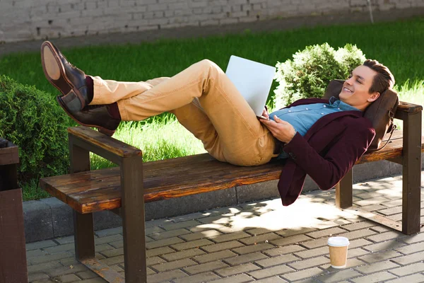 Handsome smiling young freelancer lying on bench and using laptop — Stock Photo