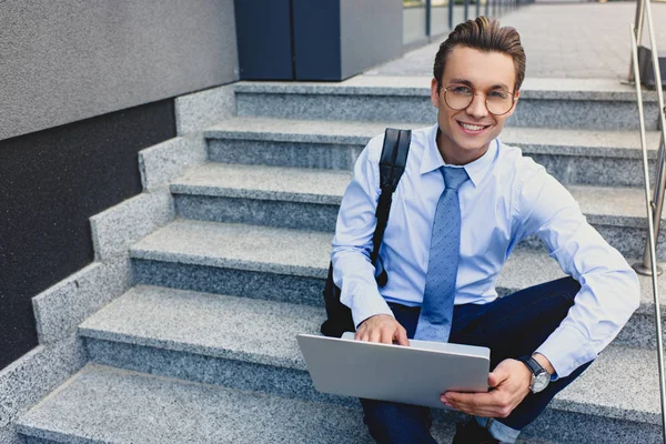 Bonito jovem empresário usando laptop e sorrindo para a câmera enquanto sentado em escadas — Fotografia de Stock