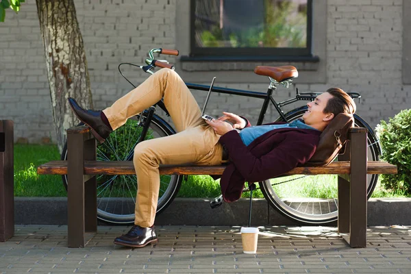 Smiling young freelancer lying on bench and using laptop — Stock Photo