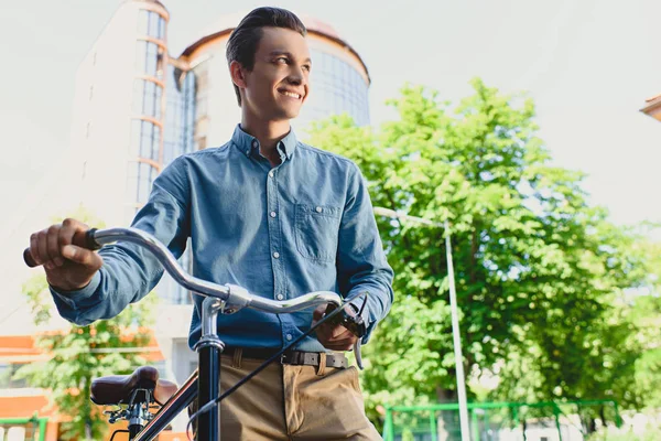 Baixo ângulo de visão de belo sorriso jovem de pé com bicicleta e olhando para longe na rua — Fotografia de Stock