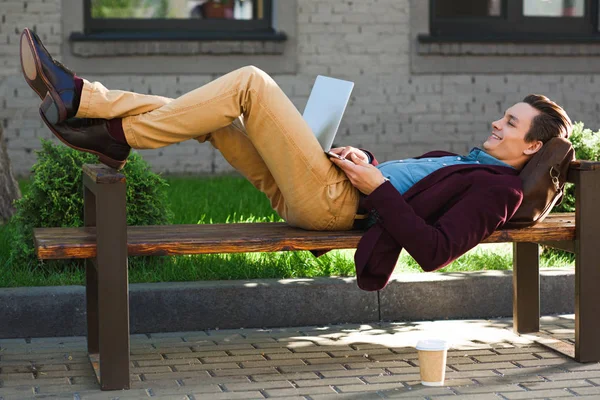 Lächelnder junger Freiberufler auf Bank liegend und mit Laptop — Stockfoto