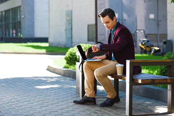 Young man using laptop and checking wristwatch while sitting on bench — Stock Photo