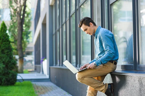 Side view of young freelancer using laptop outside modern building — Stock Photo