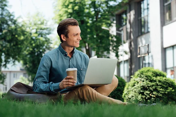 Smiling young man holding paper cup and using laptop while sitting on grass — Stock Photo