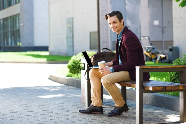 Handsome young freelancer using laptop and smiling at camera while sitting on bench — Stock Photo