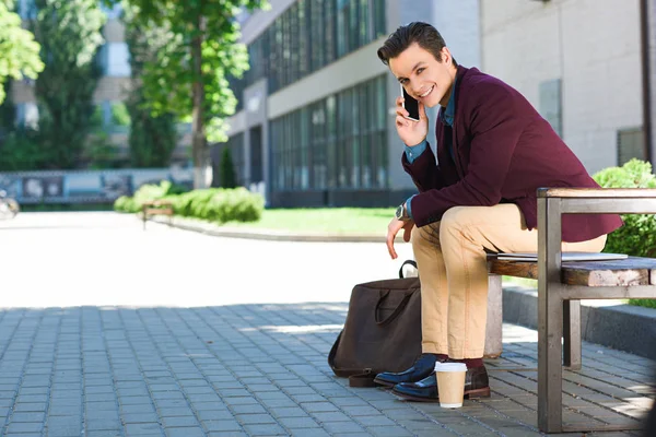 Handsome young man talking by smartphone and smiling at camera while sitting on bench — Stock Photo