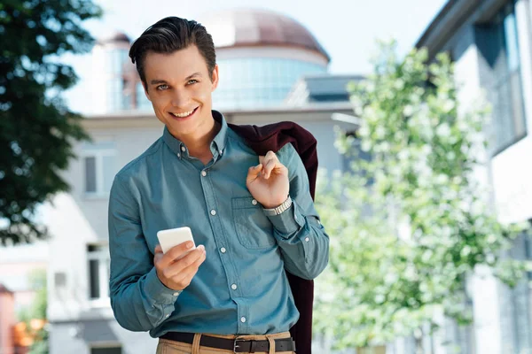 Bello giovane uomo che tiene smartphone e sorride alla fotocamera — Foto stock