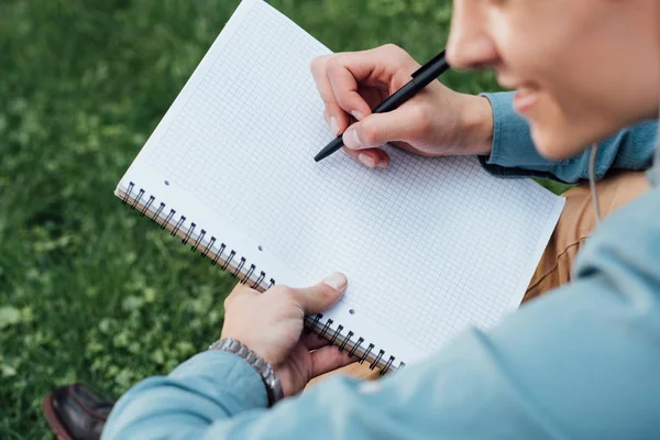 Schnappschuss eines lächelnden jungen Mannes, der im Gras sitzt und in ein leeres Notizbuch schreibt — Stockfoto
