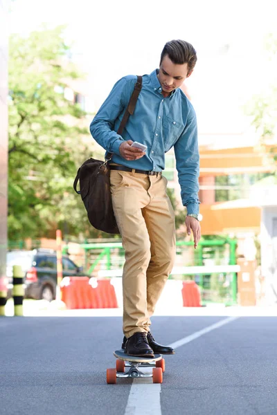 Красивый молодой человек с помощью смартфона и езды на длинной доске на улице — стоковое фото