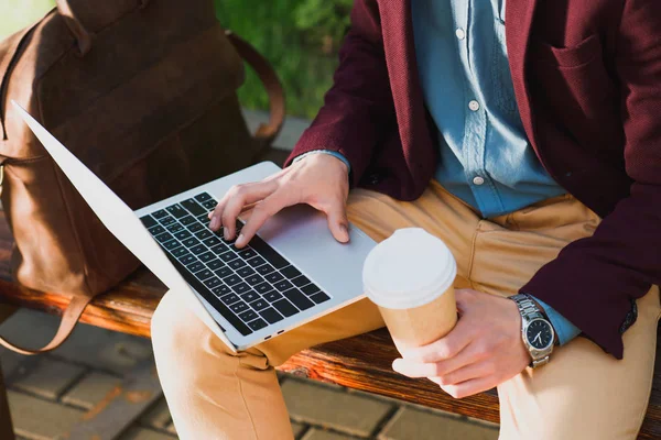 Tiro recortado de jovem freelancer segurando copo de papel e usando laptop enquanto sentado no banco — Fotografia de Stock