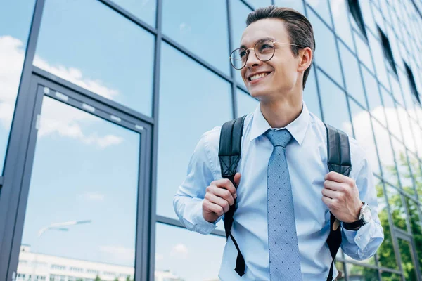 Bonito sorridente jovem empresário em óculos vestindo mochila e olhando para longe — Fotografia de Stock