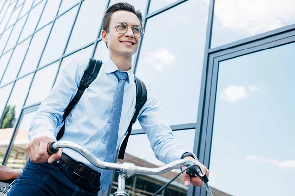 Low angle view of handsome smiling young businessman in formal wear riding bicycle and looking away — Stock Photo