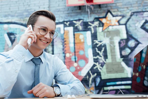 Красивый улыбающийся молодой фрилансер в очках разговаривает со смартфоном и пользуется ноутбуком — стоковое фото