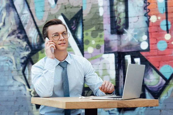 Joven guapo en gafas que habla por teléfono inteligente y el uso de ordenador portátil - foto de stock