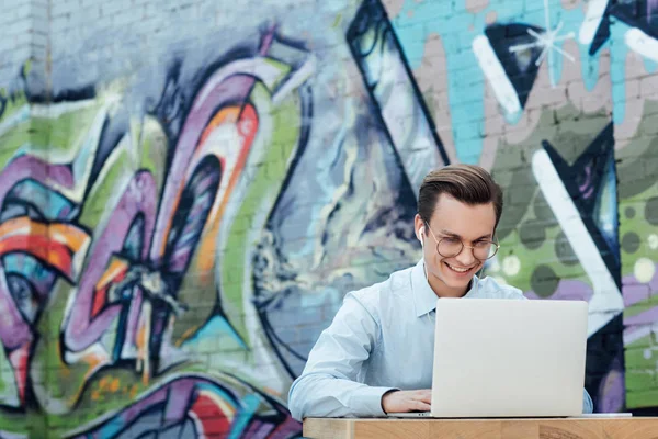 Sonriente joven freelancer en gafas usando laptop mientras está sentado afuera - foto de stock