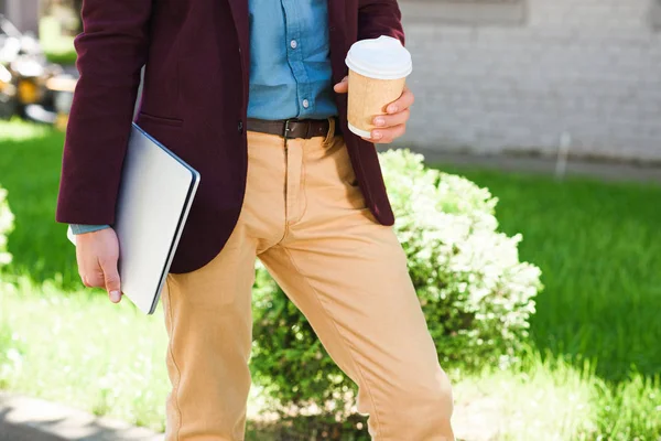 Обрезанный снимок молодого человека с ноутбуком и бумажной чашкой снаружи — стоковое фото