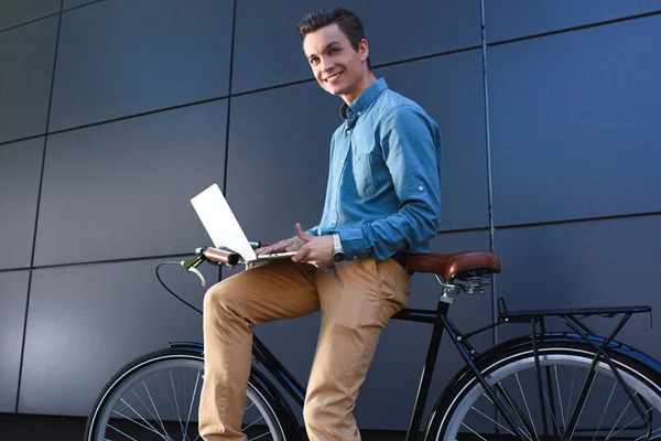 Smiling young male freelancer sitting on bike and using laptop — Stock Photo