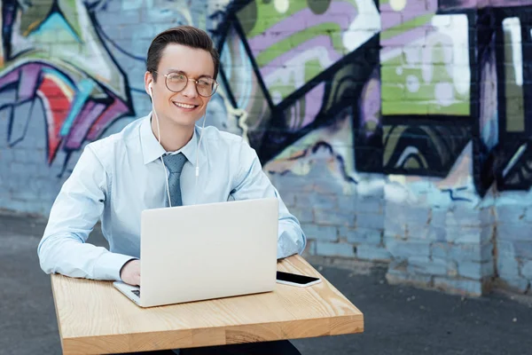 Jovem empresário bonito em óculos usando laptop e sorrindo para a câmera fora — Fotografia de Stock