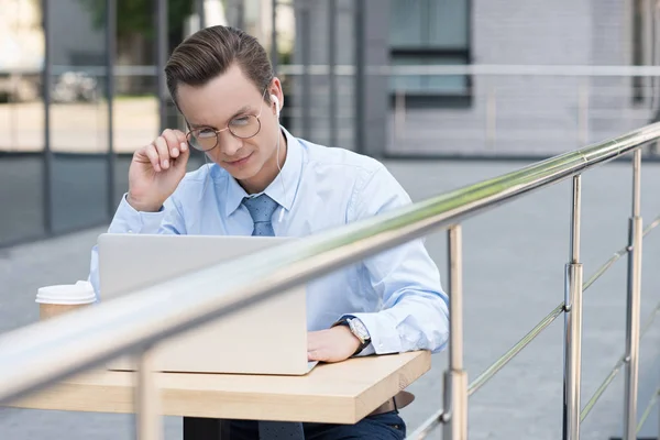 Young freelancer in eyeglasses using laptop while sitting outside modern building — Stock Photo