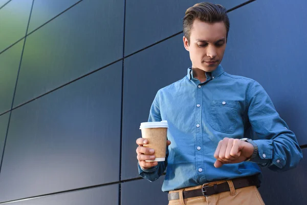 Low angle view of young man holding paper cup and checking wristwatch — Stock Photo