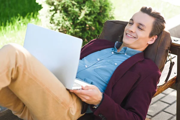 Feliz jovem freelancer deitado no banco e usando laptop — Fotografia de Stock