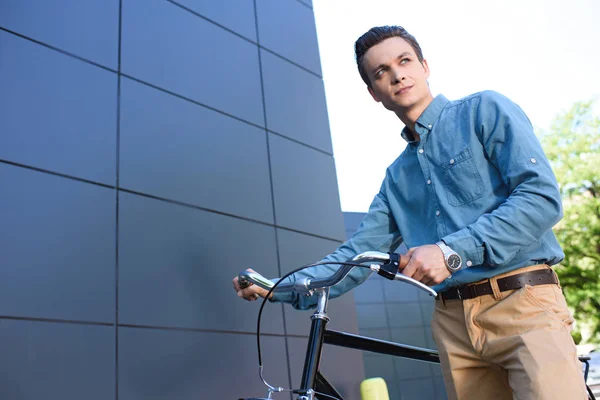 Low angle view of handsome young man standing with bike and looking away — Stock Photo