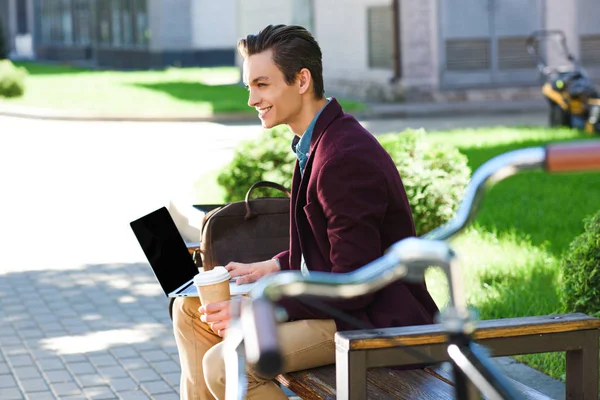 Visão lateral do jovem sorridente usando laptop com tela em branco no banco — Fotografia de Stock