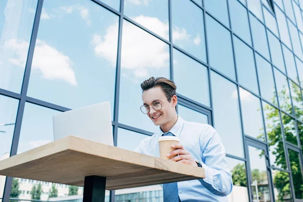 Low angle view of smiling young freelancer holding paper cup and using laptop outside modern building — Stock Photo