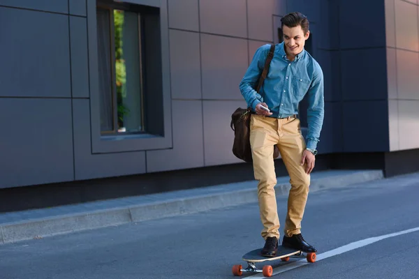 Sonriente joven montando longboard y usando smartphone - foto de stock