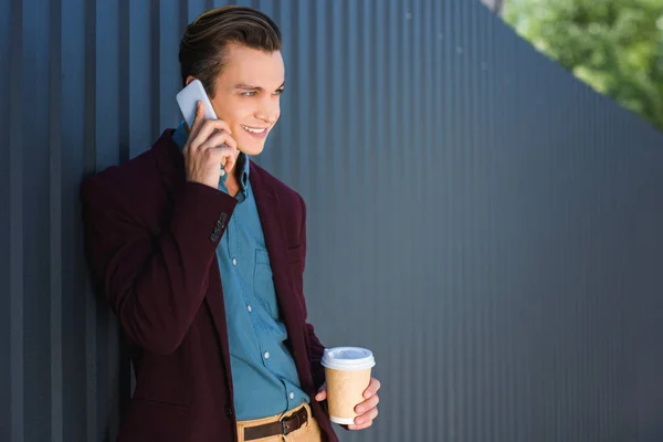 Lächelnder junger Mann hält Pappbecher in der Hand und spricht mit dem Smartphone — Stockfoto