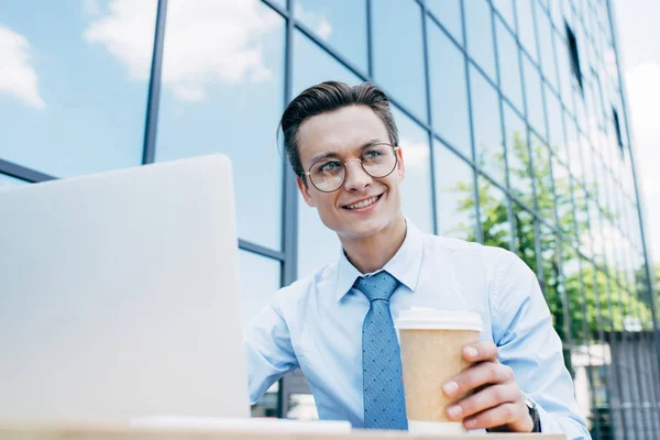 Vista angolo basso del giovane uomo d'affari sorridente utilizzando il computer portatile e tenendo tazza di carta — Foto stock