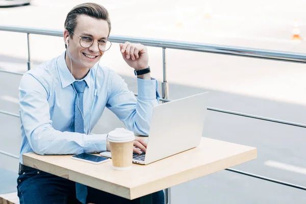 Jovem empresário sorridente em óculos usando laptop e fones de ouvido fora — Fotografia de Stock