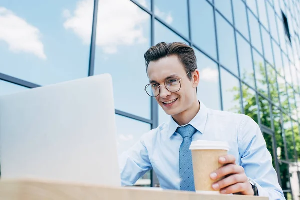 Guapo joven hombre de negocios sonriente en gafas usando el ordenador portátil y sosteniendo la taza de papel fuera del edificio moderno - foto de stock