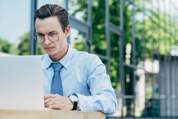 Handsome young businessman in eyeglasses using laptop outside modern building — Stock Photo