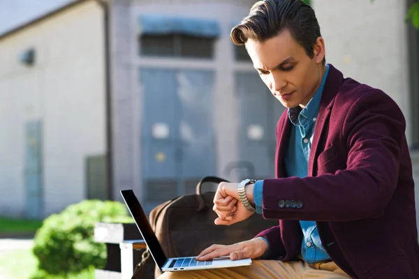 Handsome young man using laptop and checking wristwatch while sitting on bench — Stock Photo
