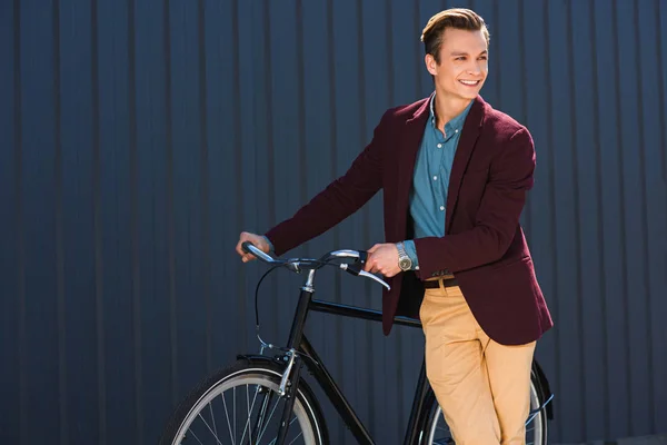 Guapo joven sonriente de pie con la bicicleta y mirando hacia otro lado - foto de stock