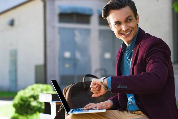 Handsome smiling young man using laptop and checking wristwatch — Stock Photo