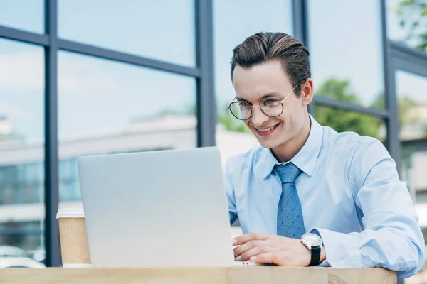Bello sorridente giovane uomo in occhiali da vista utilizzando computer portatile fuori edificio moderno — Foto stock