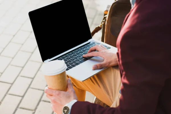 Обрізаний знімок людини, що тримає паперову чашку і використовує ноутбук з порожнім екраном на вулиці — стокове фото