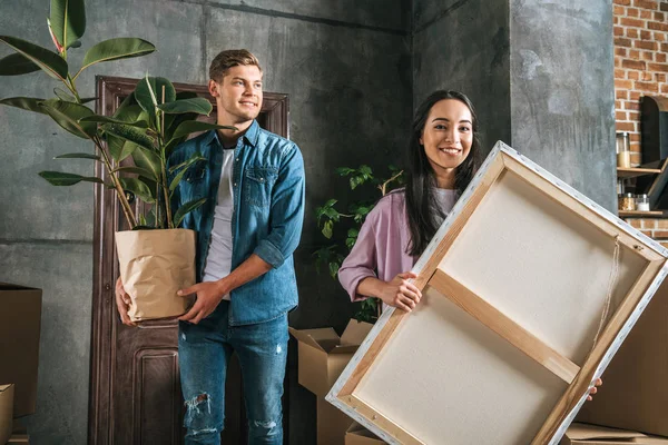 Happy young woman carrying canvas and her boyfriend holding ficus plant while moving into new home — Stock Photo