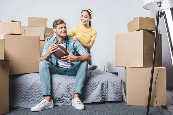 Hermosa pareja joven sentada en el dormitorio de un nuevo hogar con pelota de fútbol americano - foto de stock