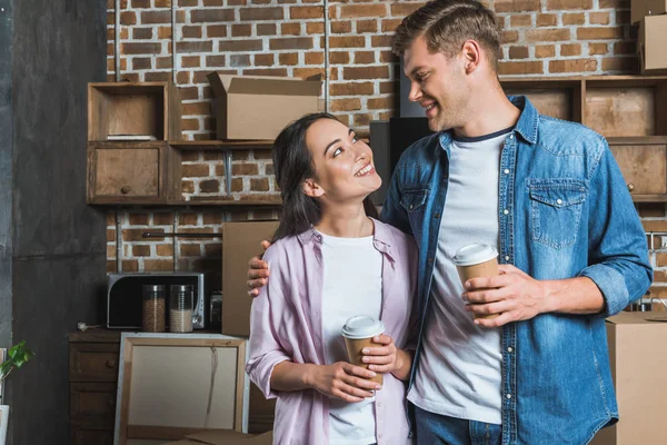 Young interracial couple with paper cups of coffee standing on kitchen while moving into new home and looking at each other — Stock Photo