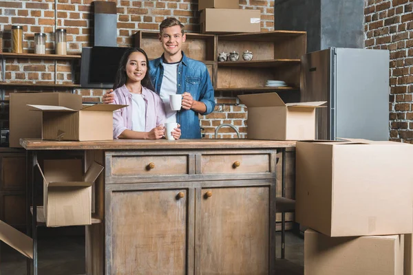 Hermosa pareja joven con tazas de café y cajas de pie en la cocina durante la mudanza a un nuevo hogar - foto de stock