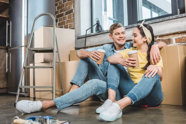 Junges gemischtrassiges Paar mit Pappbechern Kaffee auf dem Boden sitzend beim Einzug in ein neues Zuhause — Stockfoto