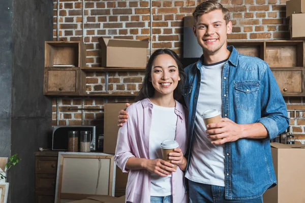 Young interracial couple with paper cups of coffee standing on kitchen while moving into new home — Stock Photo