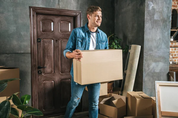 Handsome young man carrying box while moving into new house — Stock Photo