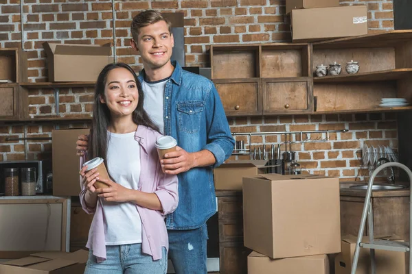 Hermosa pareja joven con tazas de papel de café de pie en la cocina mientras se muda a un nuevo hogar — Stock Photo