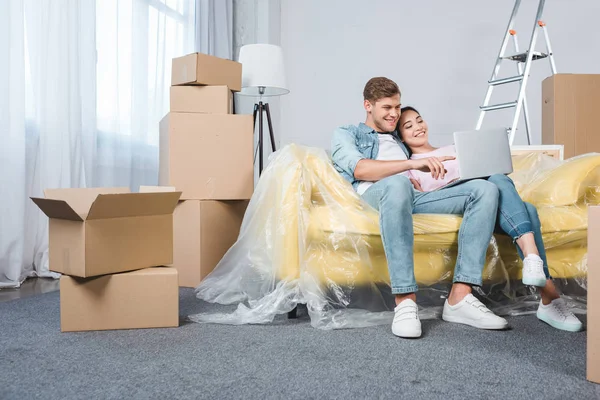 Belo jovem casal usando laptop juntos no sofá enquanto se movem para uma nova casa — Fotografia de Stock